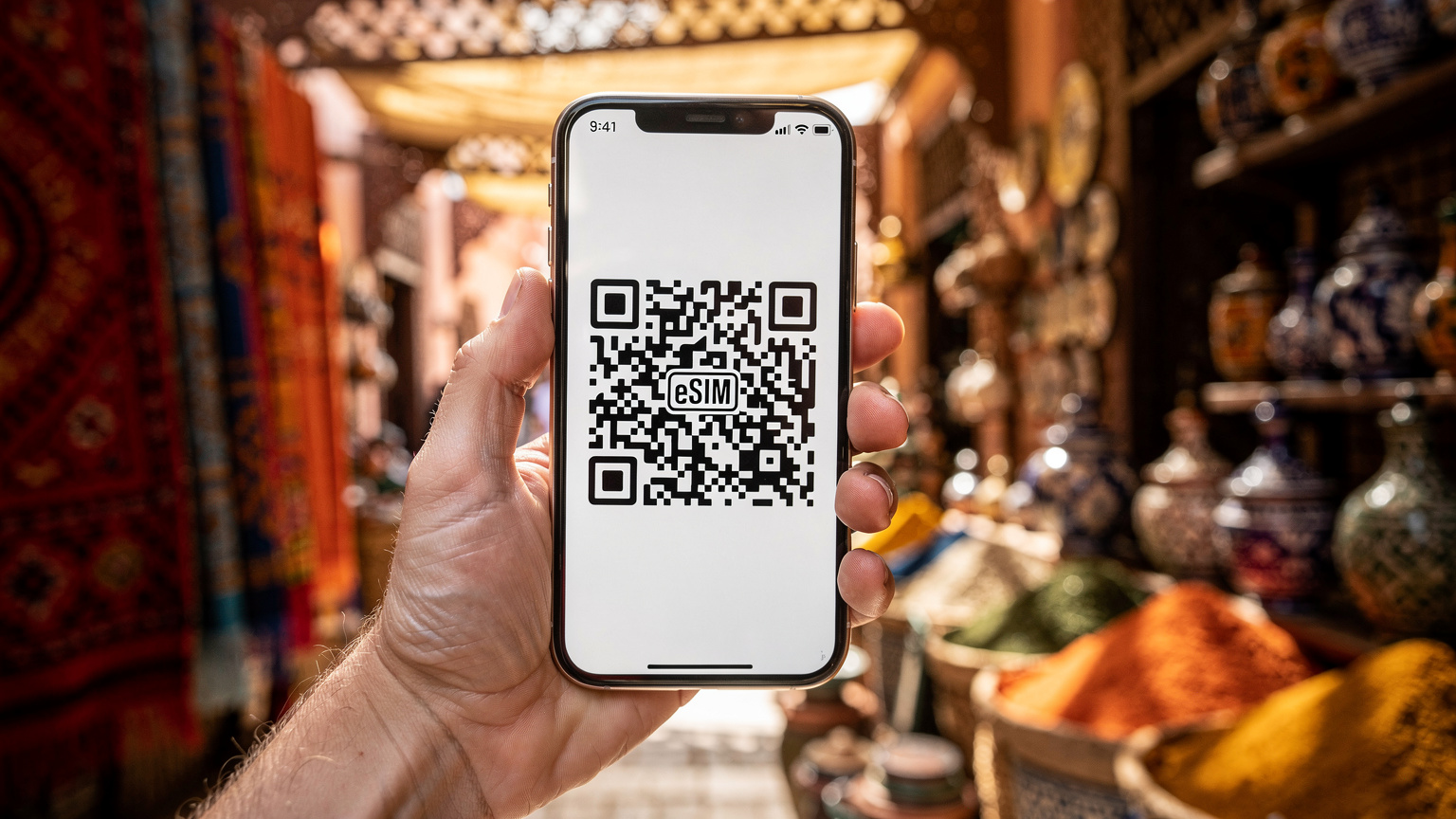 Une main tenant un smartphone affichant un code QR pour l'activation d'une eSIM, dans l'ambiance chaleureuse d'un souk marocain, avec des poteries et des tissus colorés en arrière-plan flou.