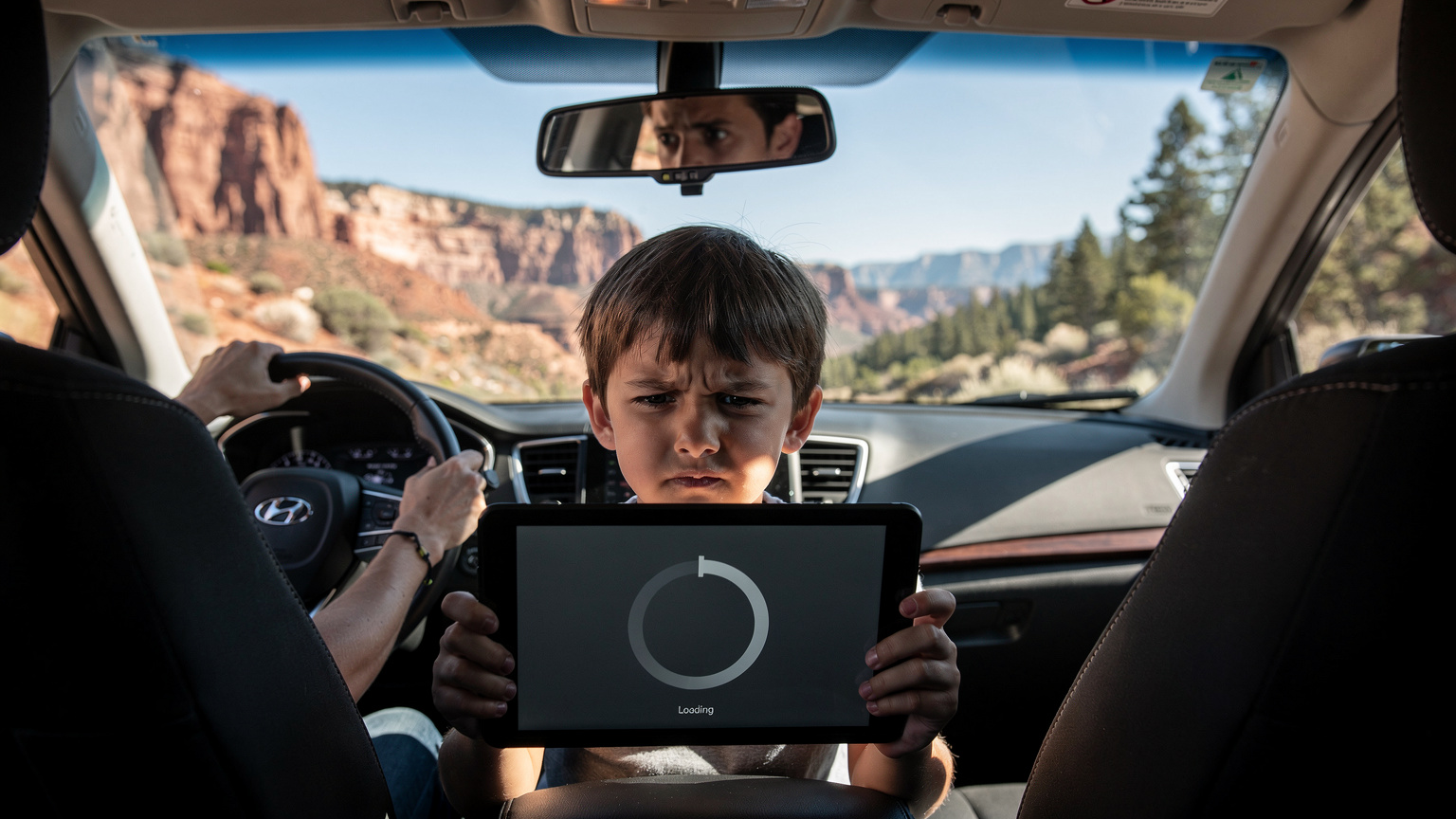 A child, aged 10, looking slightly frustrated at a buffering video on a tablet while seated in the back of a rental car, driving through a scenic but somewhat remote landscape in a US National Park, with a parent in the driver's seat glancing in the rearview mirror.