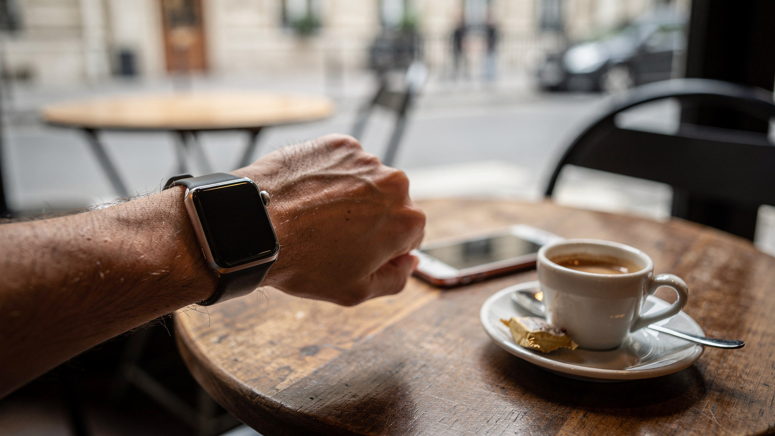 Nahaufnahme einer Hand, die auf einer Apple Watch tippt, während sie in einem belebten Café in Paris sitzt, ein iPhone liegt unscharf im Hintergrund.