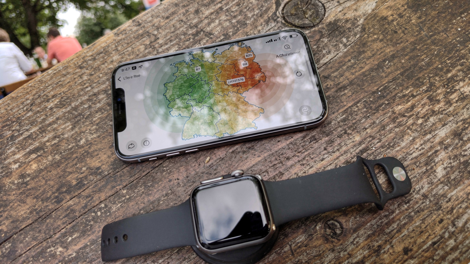Nahaufnahme eines Smartphones, das eine Karte mit eingezeichneten Frequenzbändern anzeigt, während eine Apple Watch im Vordergrund auf einem Tisch in einem Münchner Biergarten liegt.