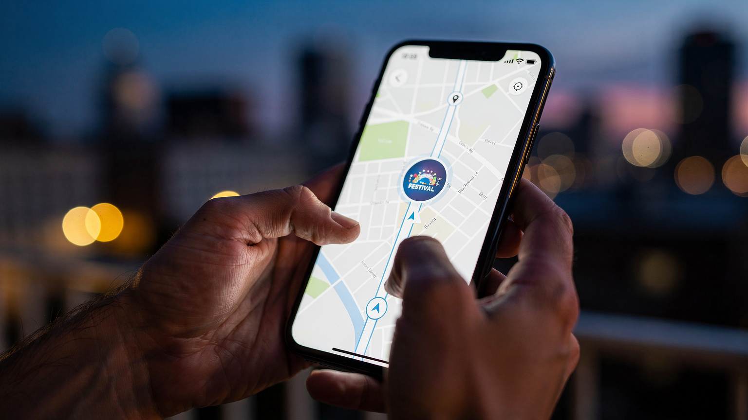 Χέρια κρατούν ένα smartphone με μια εφαρμογή χαρτών ανοιχτή, δείχνοντας μια διαδρομή προς έναν χώρο φεστιβάλ, ενώ στο φόντο διακρίνονται θολά φώτα πόλης.