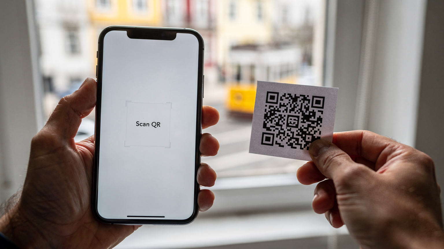 Detailní záběr rukou cestovatele, který v Lisabonu naskenuje QR kód pro aktivaci eSIM na moderním smartphonu, v pozadí je rozmazané okno s výhledem na město.