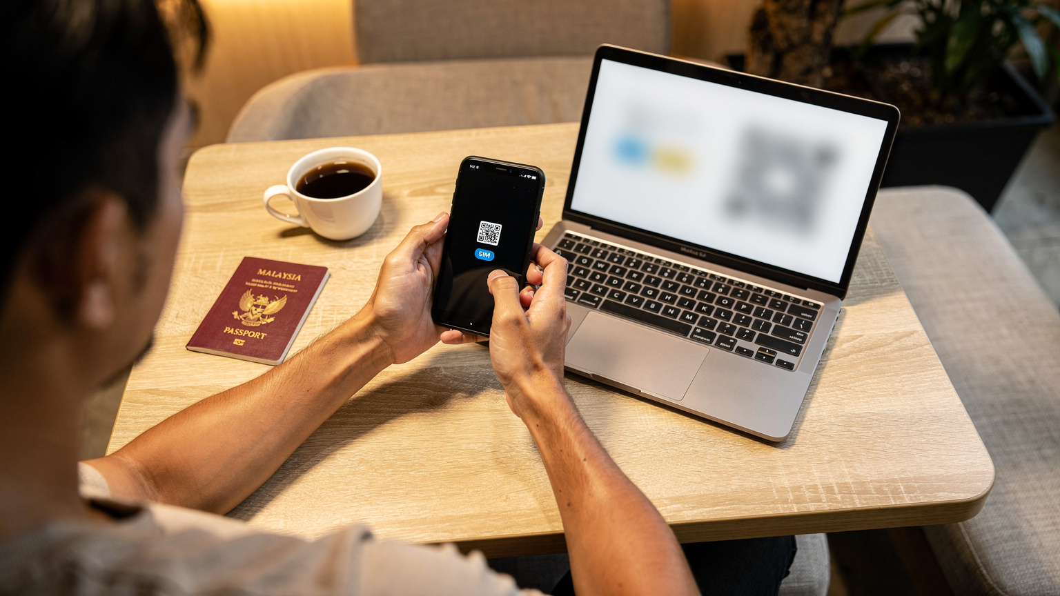 Seorang lelaki Malaysia sedang mengimbas kod QR eSIM dari skrin komputer riba di meja kafe yang santai, dengan pasport Malaysia dan secawan kopi di sebelahnya.