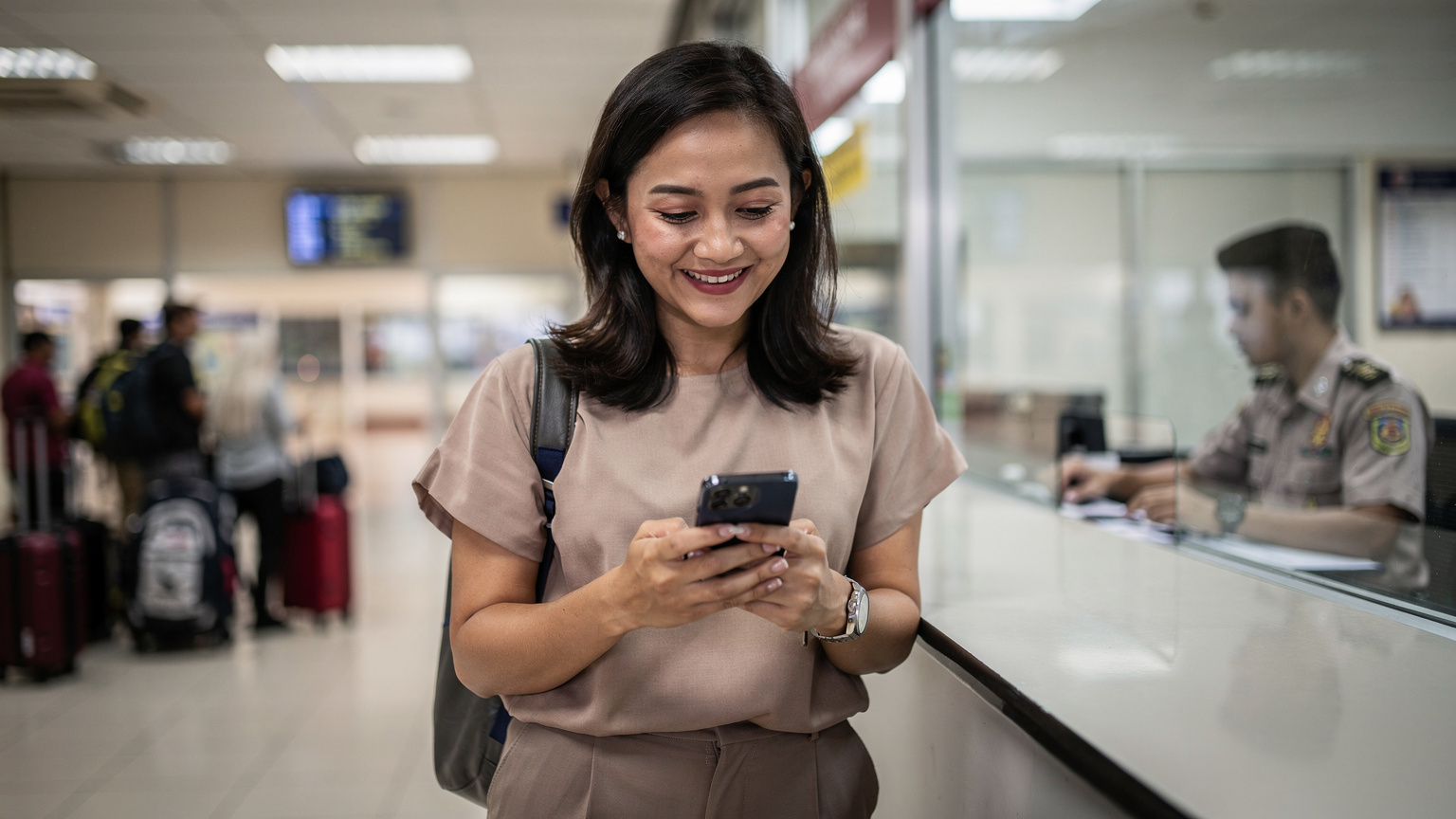 Seorang pengembara wanita tersenyum lega sambil memegang telefon bimbitnya di kaunter imigresen sempadan darat Thailand-Malaysia, dengan petugas imigresen yang kabur di latar belakang.