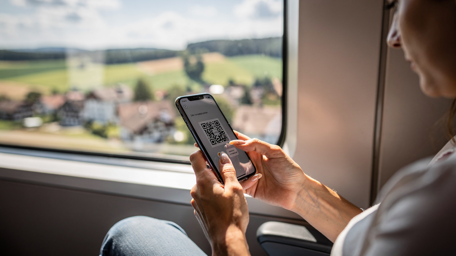 Українка легко сканує QR-код eSIM на своєму iPhone, сидячи в сучасному потязі ICE, який проїжджає по мальовничому німецькому ландшафту.