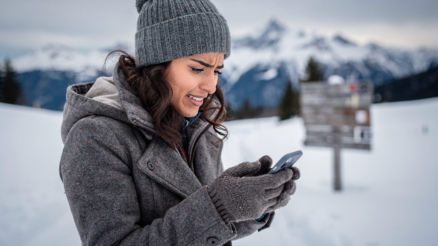 Uma mulher brasileira com um casaco de inverno elegante e um chapéu de lã, olha para o telefone com uma expressão de preocupação e um sorriso tenso, tentando encontrar sinal em uma área montanhosa da Áustria, com a neve e uma placa de trilha ao fundo.