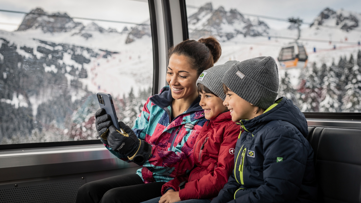 Uma mãe brasileira, sorrindo, segura seu smartphone em uma luva de esqui e mostra para seus dois filhos um mapa digital de pistas enquanto estão sentados em um teleférico nos Alpes austríacos, com a neve e montanhas ao fundo.