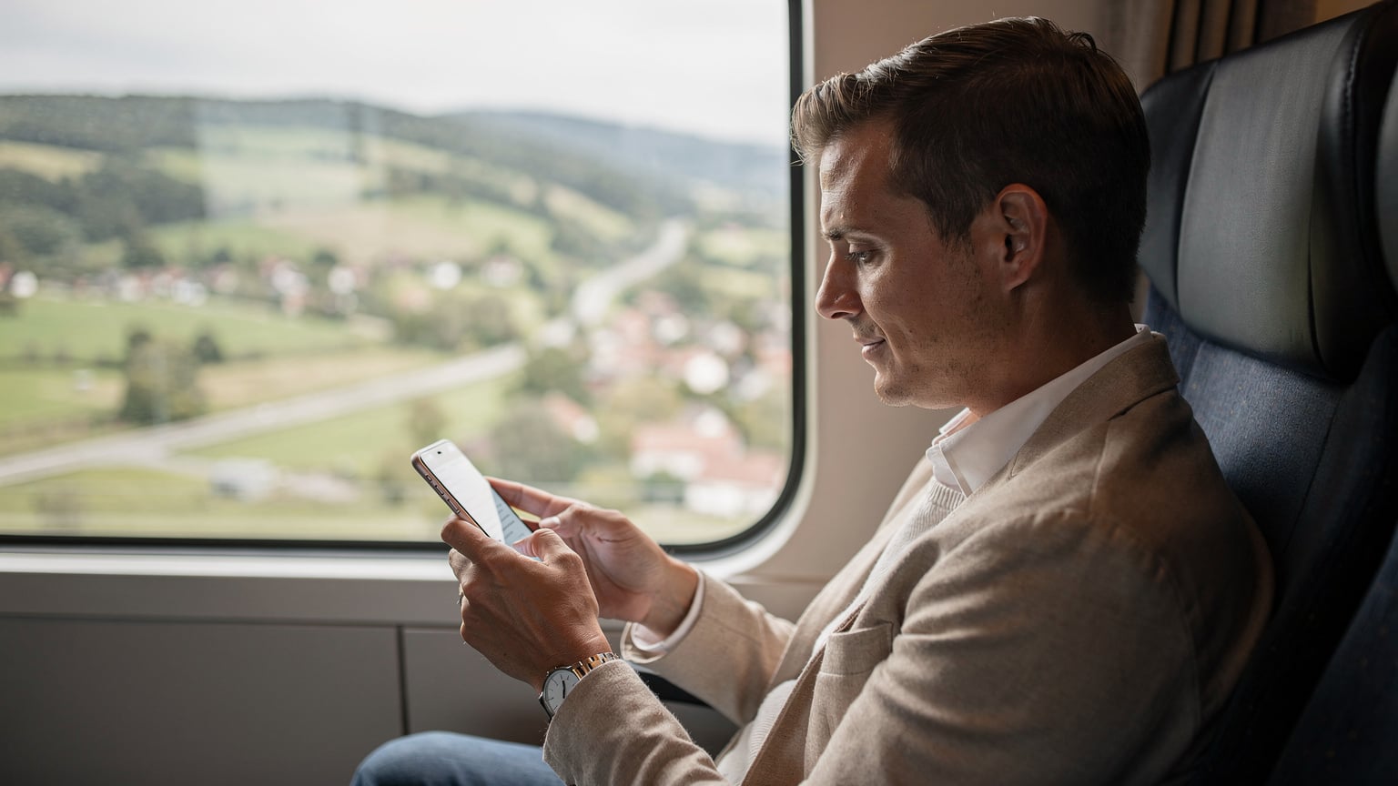 ヨーロッパの鉄道の窓から景色を眺めながら、スマホで次の駅の情報を確認している旅行者。車窓には美しい田園風景が広がる。
