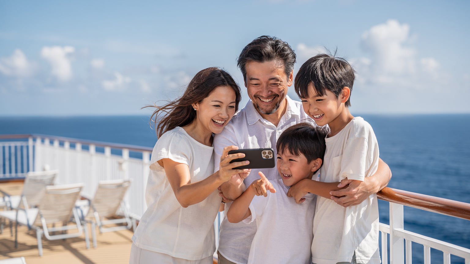 家族がクルーズ船のデッキで、海を眺めながらスマートフォンで撮影した写真を共有している。背景には青い海が広がっている。