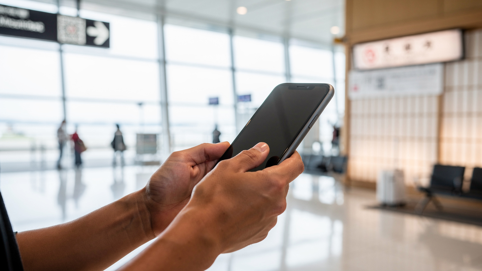 A traveler holding a smartphone, scanning a QR code for eSIM activation against the blurred background of a modern airport terminal in Japan.