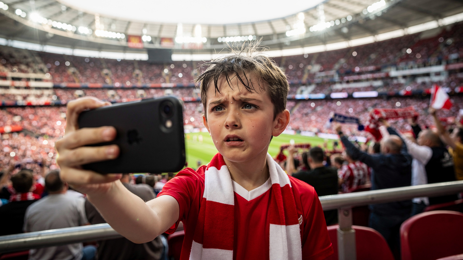 Ein junger Fußballfan im Wembley-Stadion versucht, ein Selfie zu machen, während sein Handy Schwierigkeiten hat, eine Verbindung herzustellen, im Hintergrund jubelnde Massen.