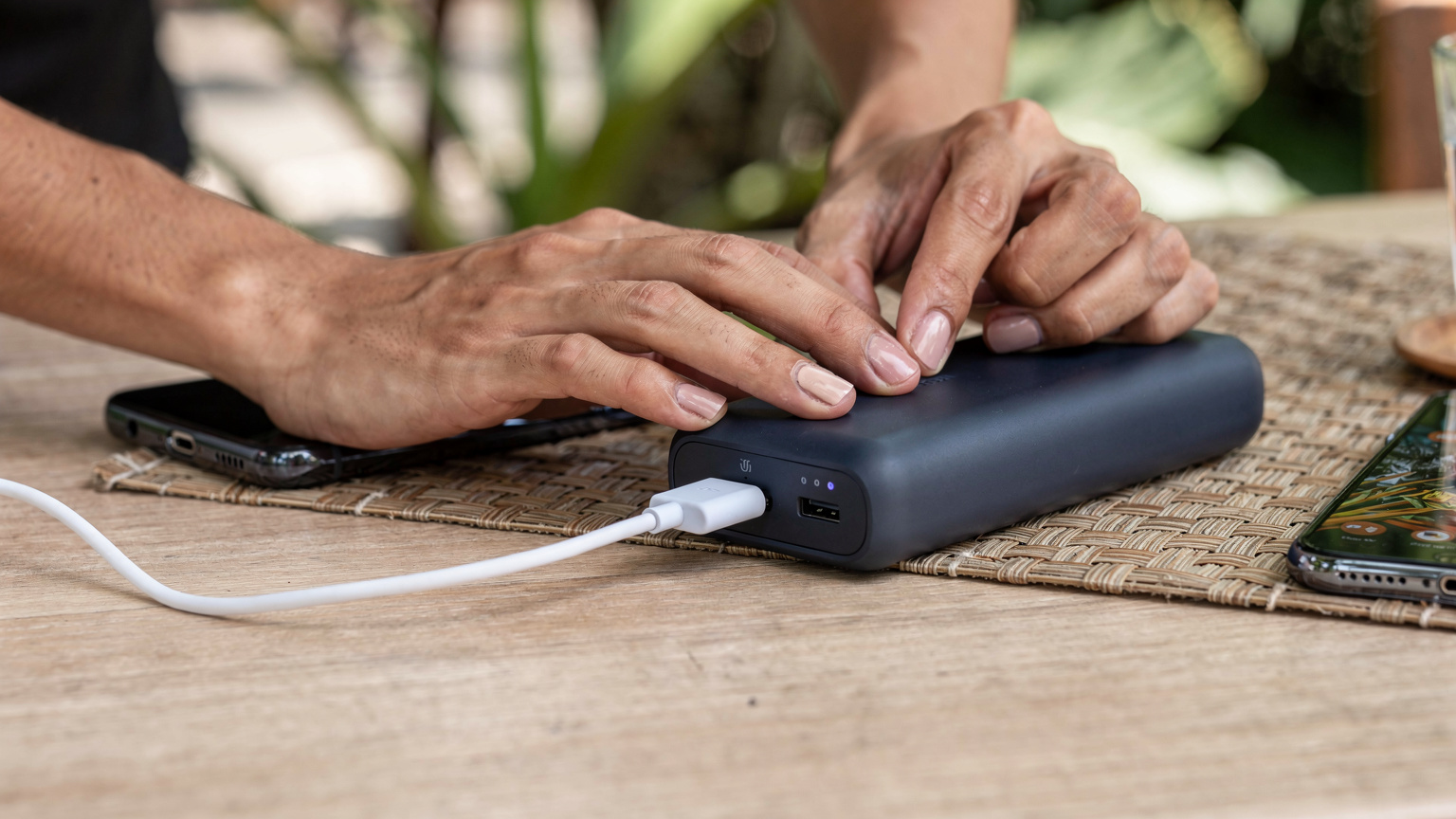 Tangan seorang wisatawan sedang menyambungkan kabel pengisi daya ke power bank berwarna biru tua, sementara di sebelahnya terlihat ponsel lain yang terhubung ke hotspot.