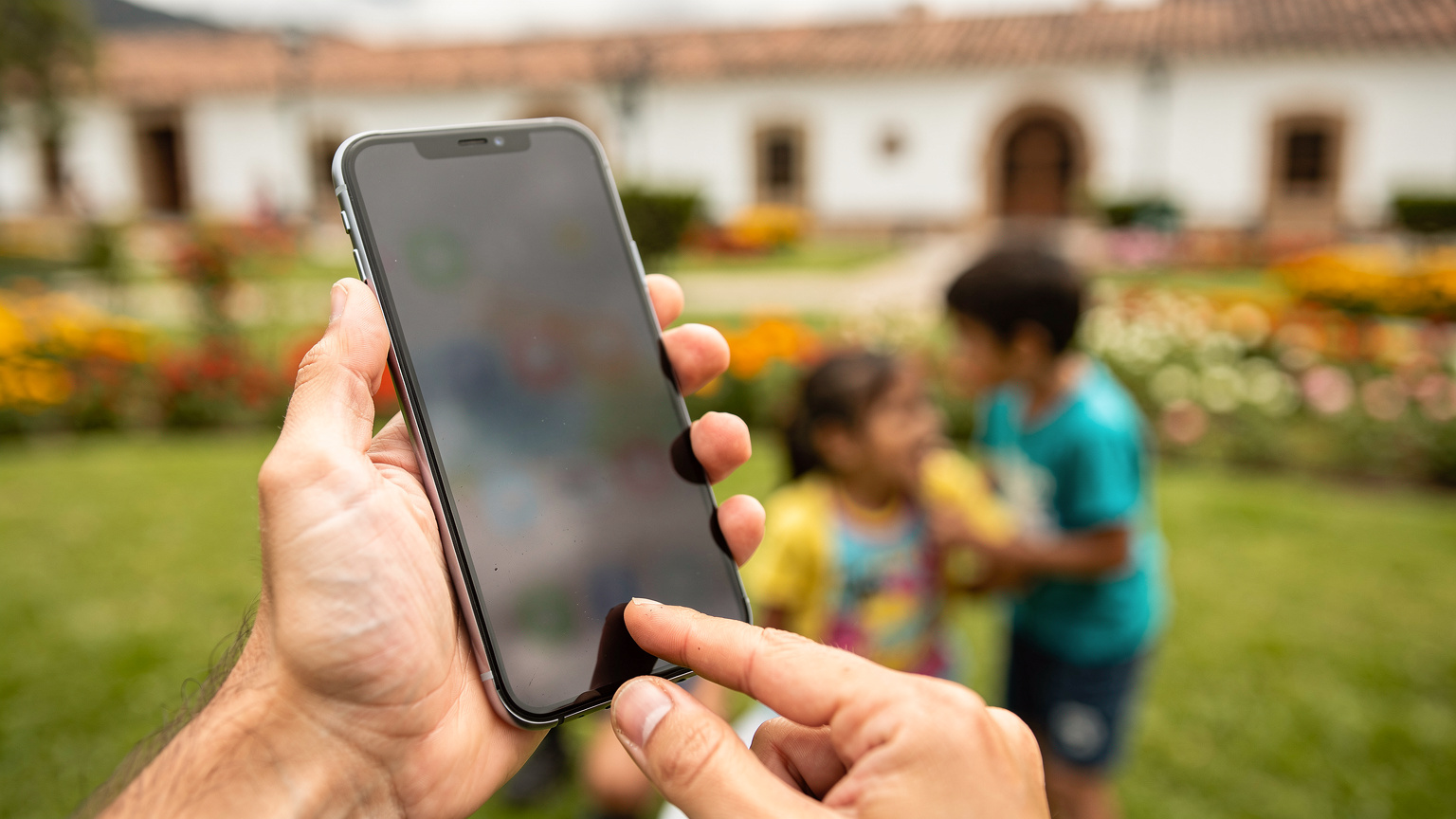 Mãos de um pai ou mãe usando um aplicativo de controle parental em um smartphone, com crianças brincando em um parque vibrante ao fundo em Cuenca, Equador.