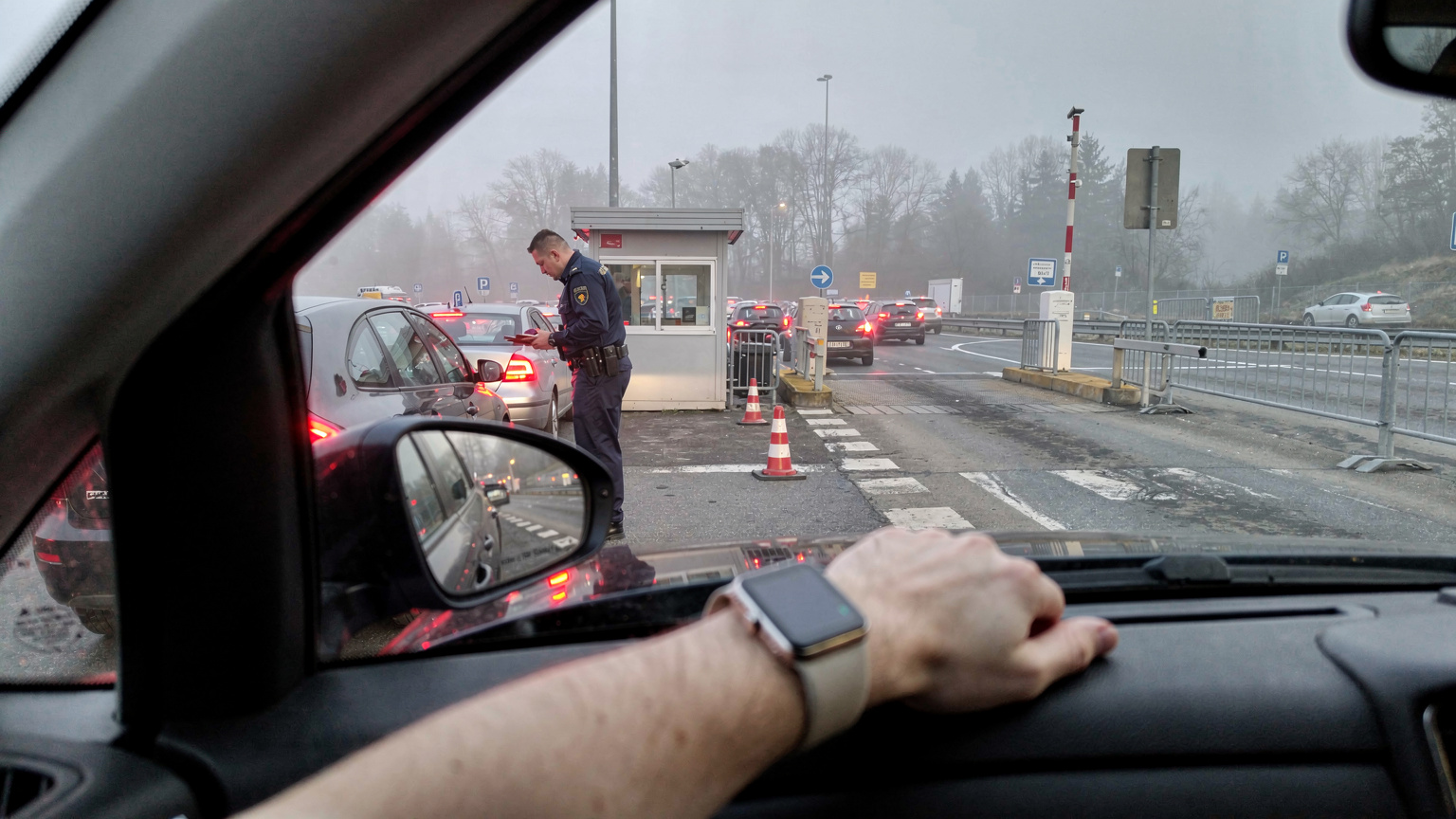 En travel grenseovergang mellom to EU-land, med biler i kø og en grensevakt som kontrollerer pass, sett fra en bil. En Apple Watch er synlig på passasjerens håndledd.