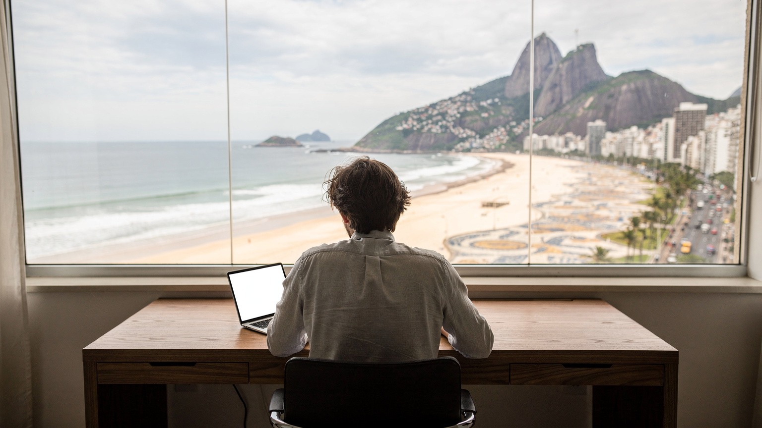 Eine Person blickt von einem Co-Working-Space am Strand in Copacabana, Rio de Janeiro, auf die belebte Promenade und den Zuckerhut in der Ferne, während sie einen Laptop auf dem Tisch vor sich hat.