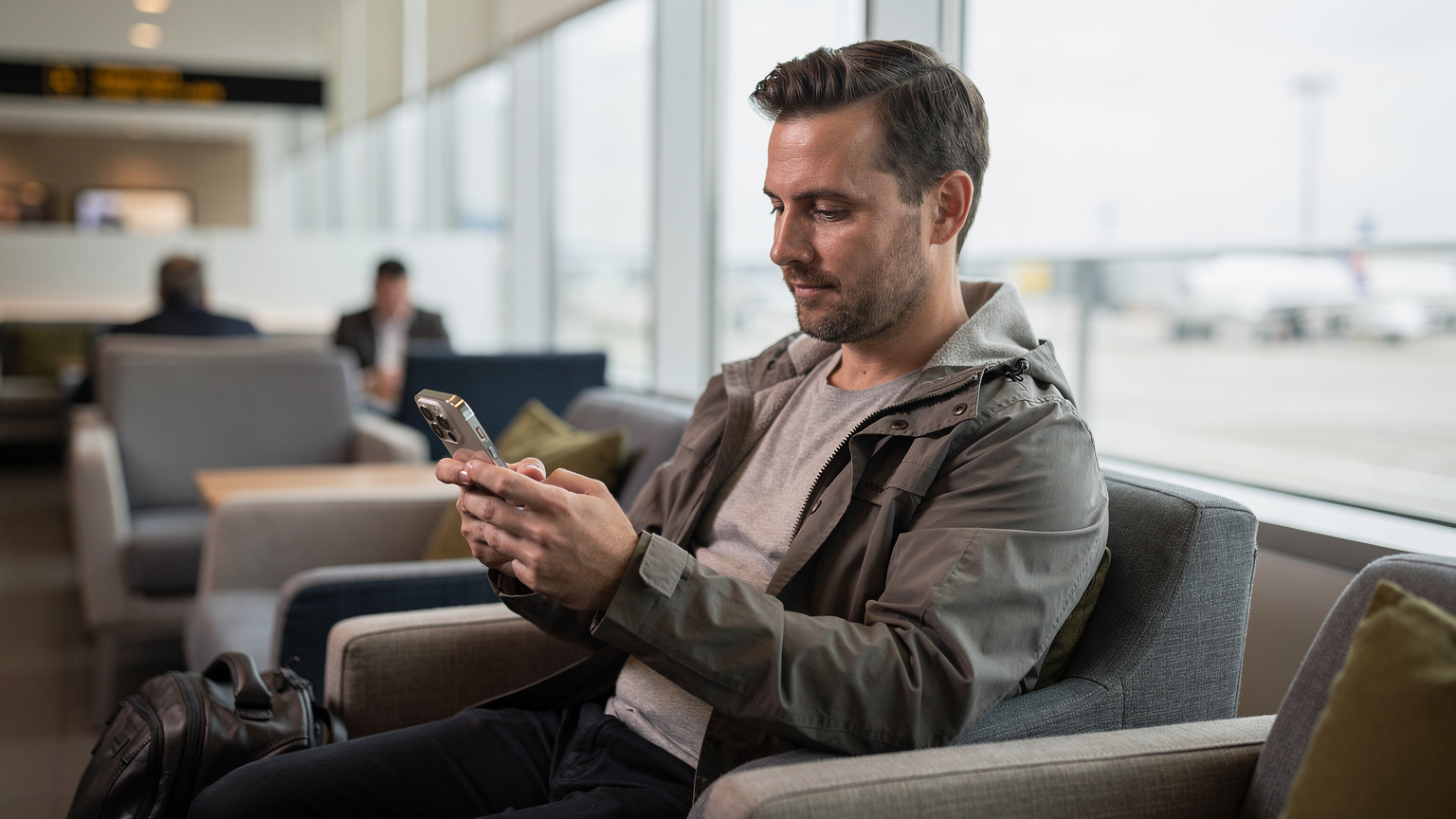 Un viajero consulta su iPhone 15 Pro, mostrando una conexión de red activa, mientras está sentado en la sala de espera del aeropuerto de Heathrow, planificando su próximo tramo de viaje.