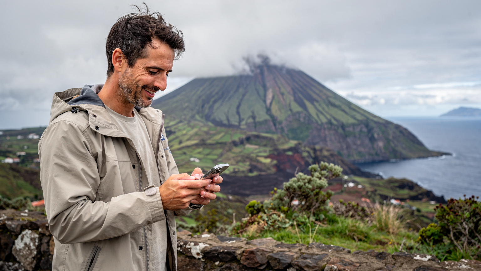 Um viajante sorri enquanto usa seu iPhone 18 para navegar por um mapa digital dos Açores, com as dramáticas montanhas vulcânicas da Ilha do Pico e o oceano Atlântico ao fundo, num dia parcialmente nublado.