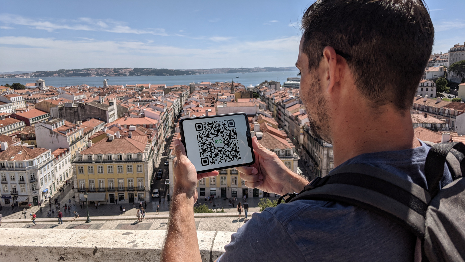 Um mochileiro, de costas, segura seu iPhone 18 com o ecrã a exibir um QR code de ativação de eSIM, enquanto observa a paisagem da Baixa Pombalina em Lisboa, com os seus edifícios característicos e o rio Tejo ao fundo, num dia de sol.