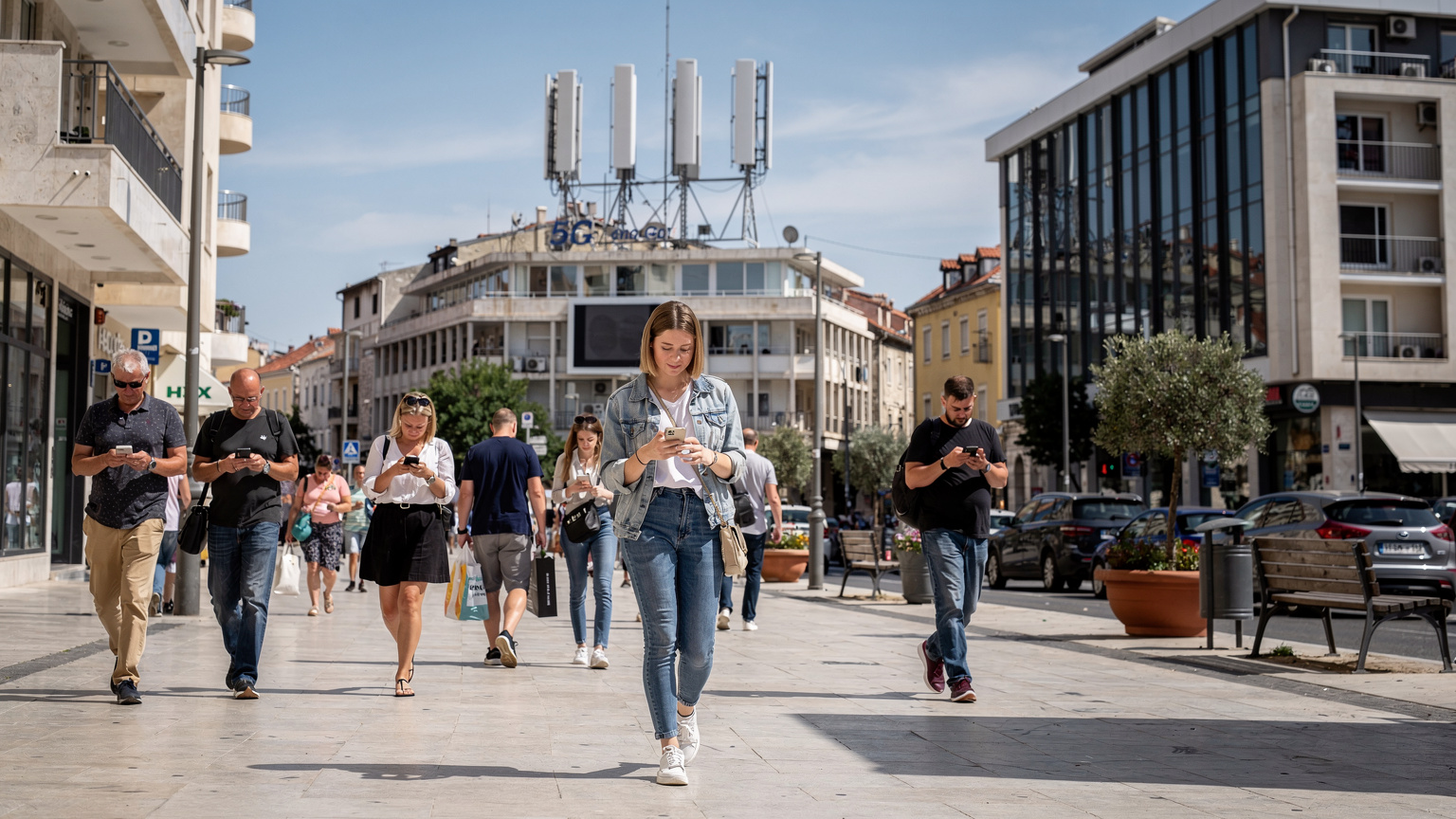 Сучасна міська вулиця в Спліті, Хорватія, з високою антеною 5G на задньому плані та людьми, які користуються смартфонами на передньому плані, що підкреслює інтеграцію технологій у повсякденне життя.