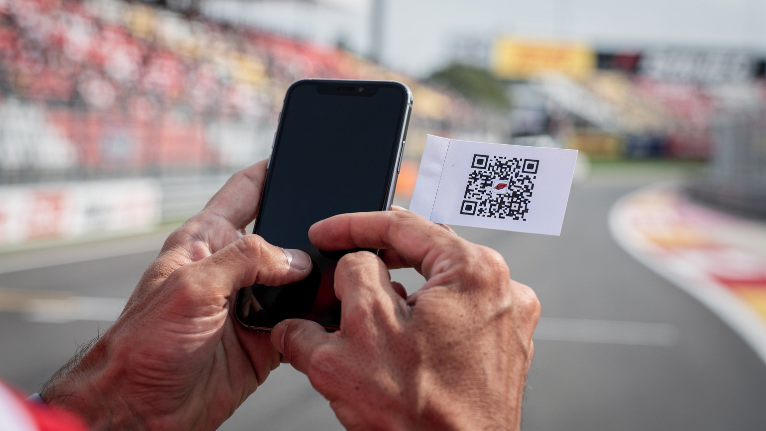 Een close-up van handen die een smartphone vasthouden, bezig met het installeren van een eSIM via een QR-code, met de wazige achtergrond van een F1-circuit op de racedag.