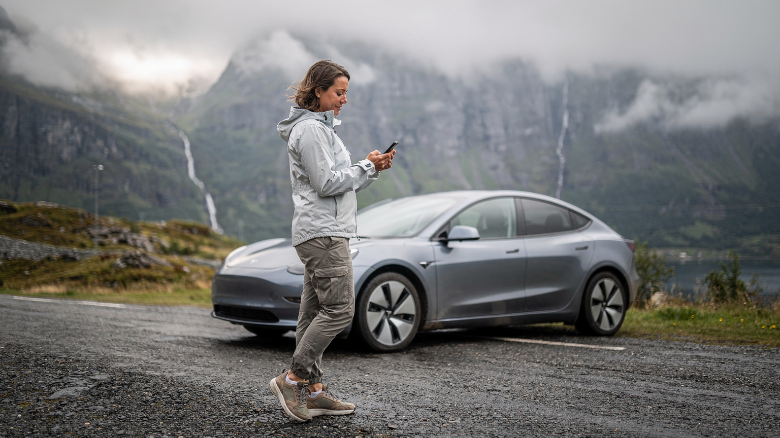 En kvinde tjekker vejret på sin smartphone ved en elbil på en grusvej i de norske fjelde, forbereder sig på vandretur.