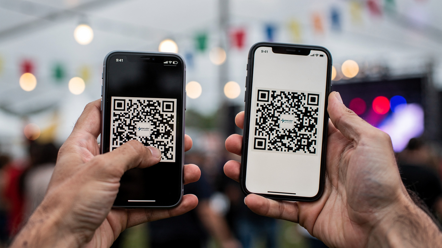 Nærbillede af hænder, der holder en smartphone og scanner en QR-kode fra en anden telefon for at aktivere et eSIM, med en baggrund af en festivalplads i et sløret fokus.