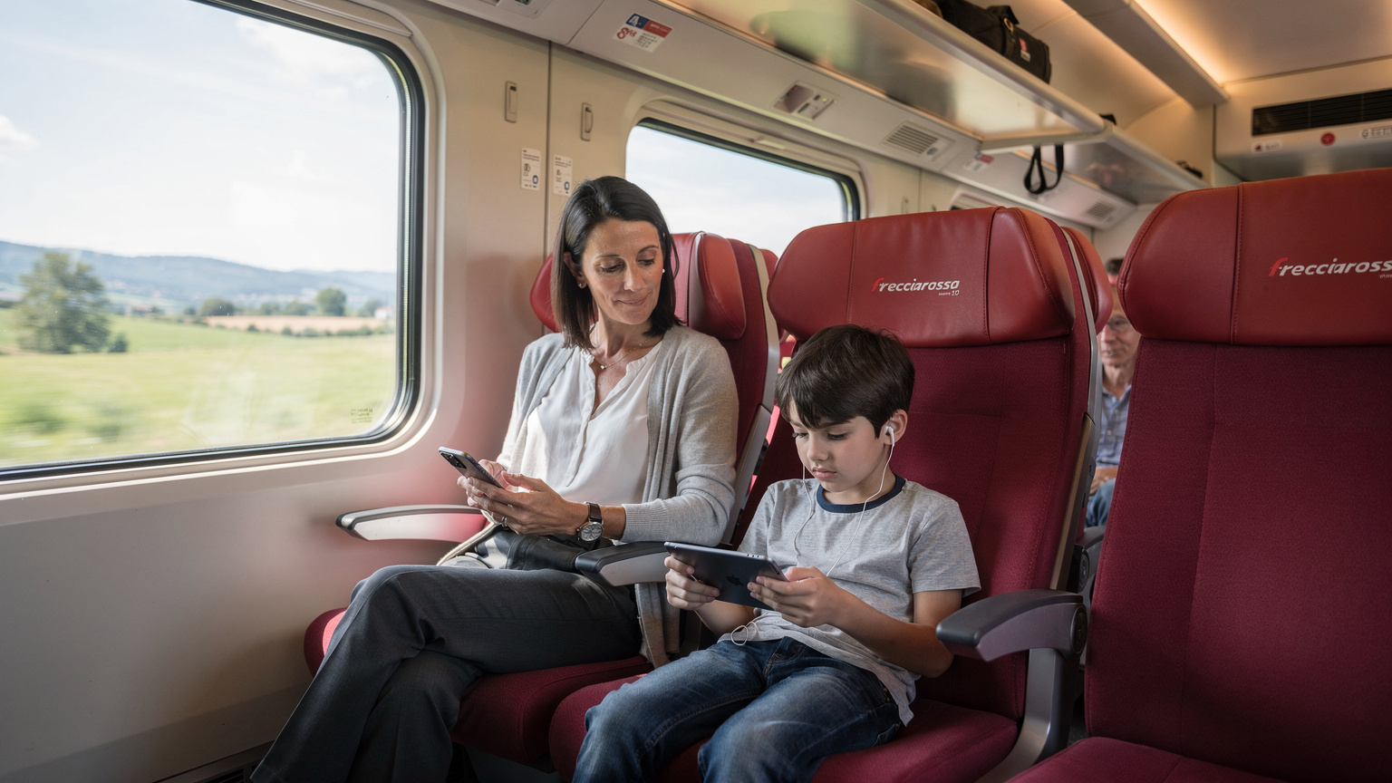 Un bambino di 8 anni è seduto su un sedile del treno ad alta velocità Frecciarossa, completamente assorto in un gioco sull'iPad, mentre la madre di fianco controlla la connessione eSIM sul suo smartphone.