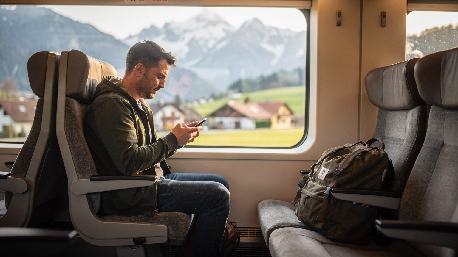 Un homme voyageur, sac à dos à ses pieds, consultant son téléphone portable depuis le siège d'un train à travers une fenêtre, avec un paysage urbain et montagneux flou en arrière-plan, sous une lumière douce du matin.