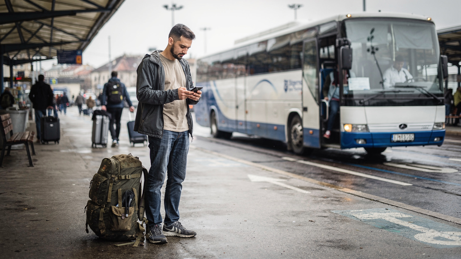 Ein junger Reisender überprüft sein iPhone an einem belebten Busbahnhof in Belgrad, sein Rucksack liegt neben ihm, während im Hintergrund ein Fernbus auf die Abfahrt wartet.
