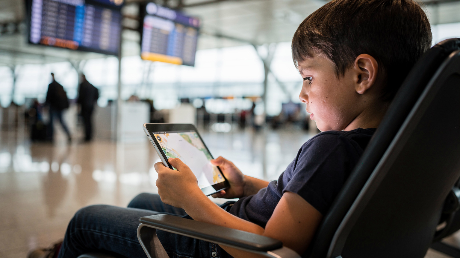 Ein Kind spielt konzentriert auf einem Tablet, während im Hintergrund verschwommen ein Flughafen-Gate zu sehen ist, was die Notwendigkeit von Unterhaltung unterwegs unterstreicht.