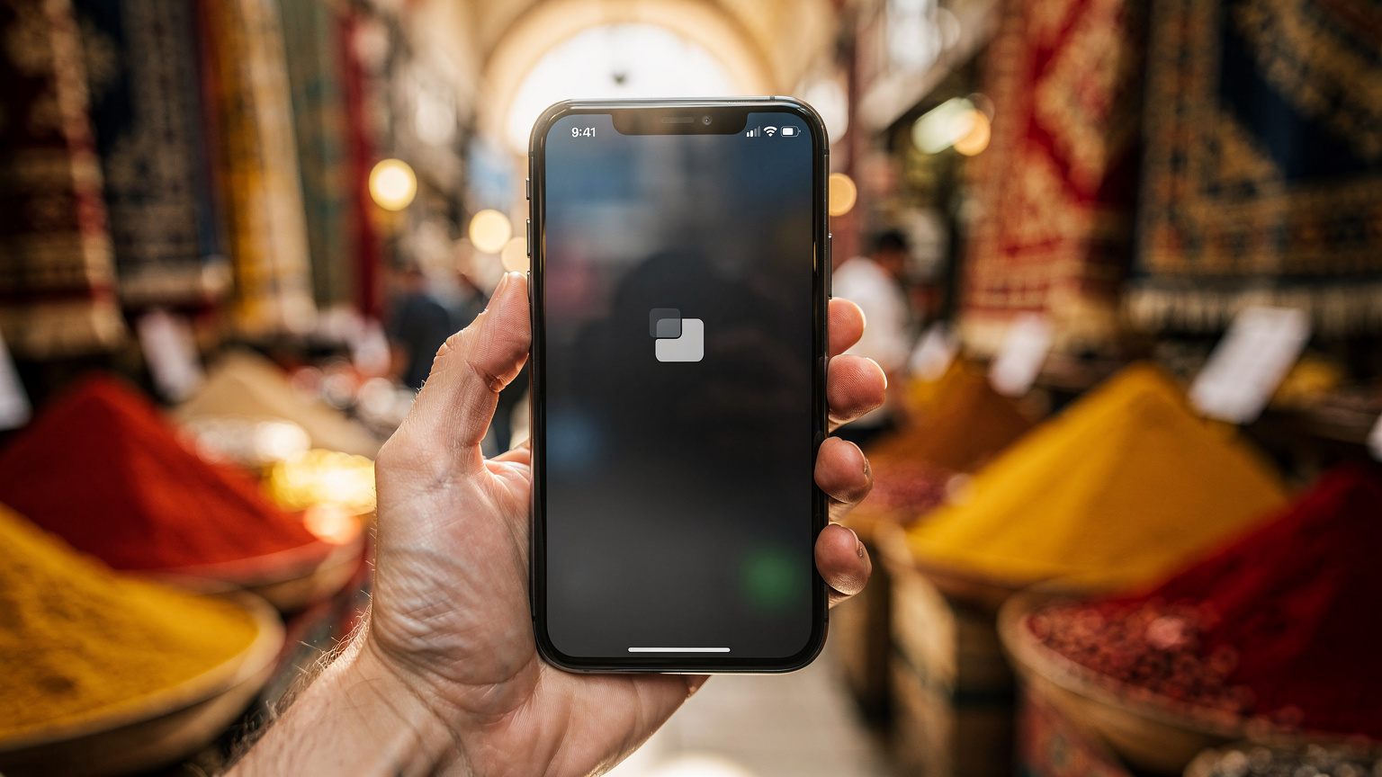 Detailný záber na ruku držiacej telefón s otvorenou eSIM aplikáciou, v pozadí je rozmazaný rušný Grand Bazaar v Istanbule s koreninami a textíliami.
