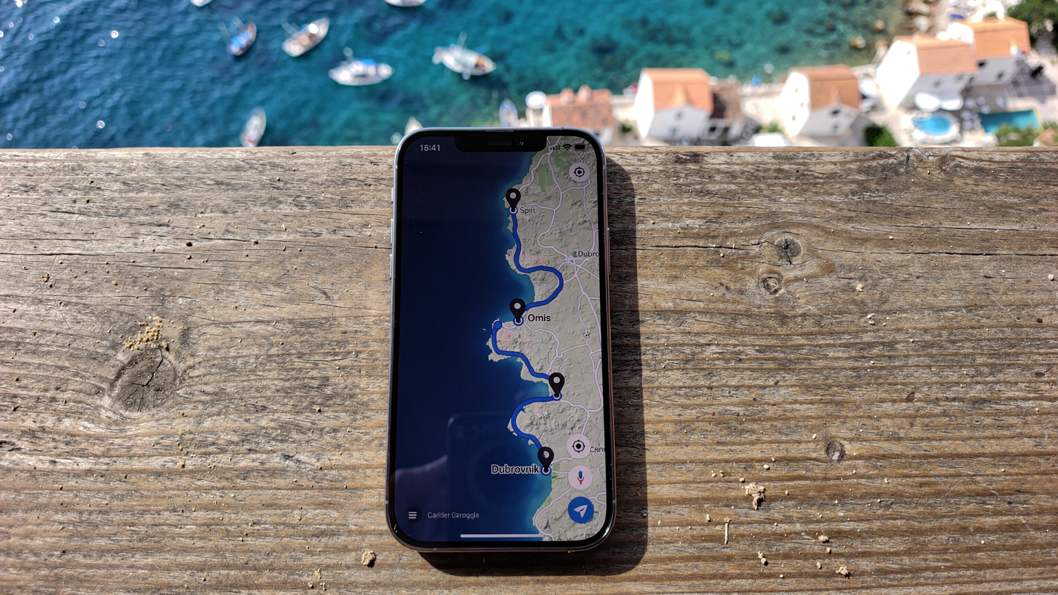 Ένα smartphone δείχνει μια εφαρμογή χαρτών με μια διαδρομή σχεδιασμένη κατά μήκος της Δαλματικής ακτής, ενώ βρίσκεται πάνω σε ένα τραπέζι καφέ με φόντο τη θάλασσα και ένα κροατικό ψαροχώρι.