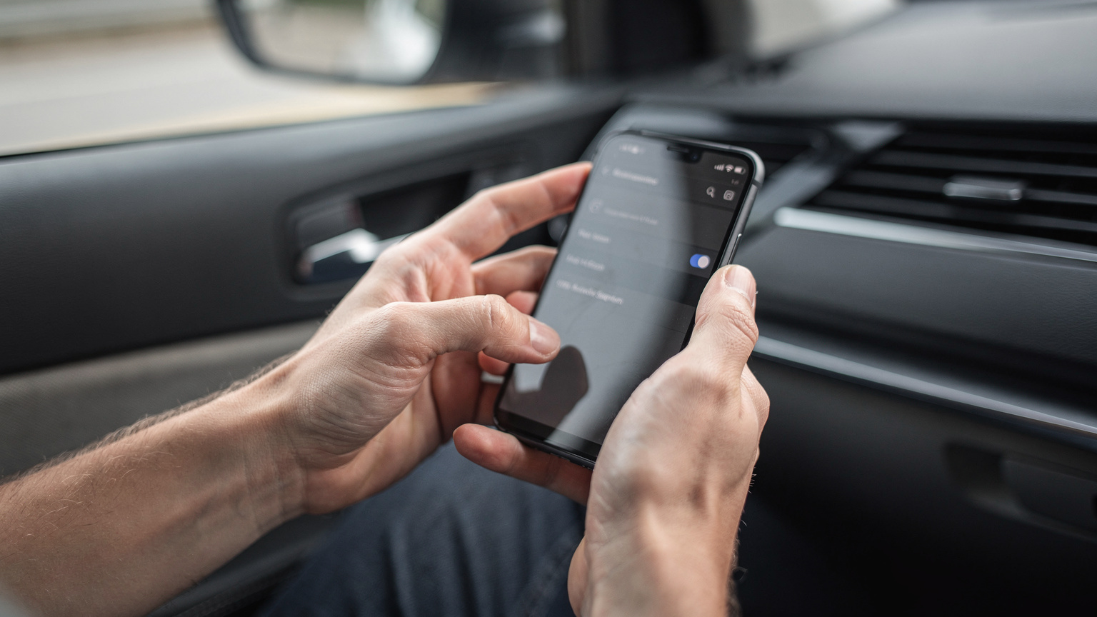 Close-up shot of a pair of hands holding a smartphone, with thumbs navigating through settings to activate an eSIM, against a blurred background of a car interior.
