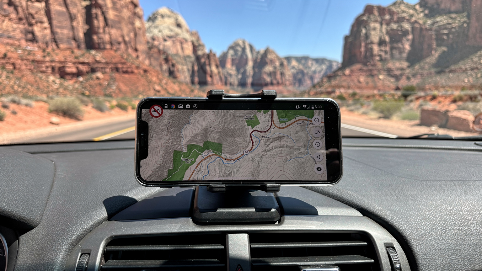 A smartphone screen displays a detailed offline map of Zion National Park, with a 'no signal' icon prominently overlaid, on a dashboard overlooking a scenic desert road.