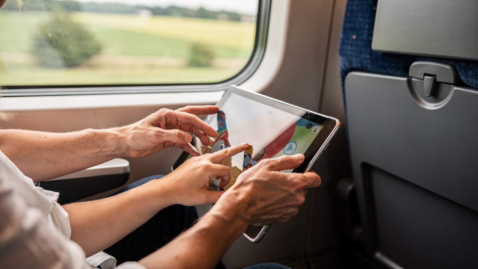 A close-up of a parent's hands gently guiding a child's fingers on a tablet screen, showing interaction with an educational game or video during a quiet moment on a train journey through the German countryside.
