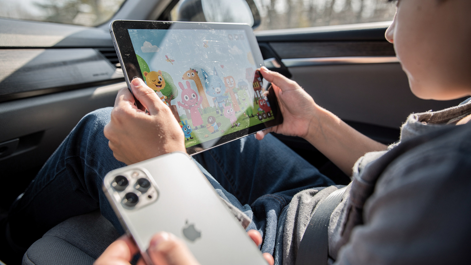 A child's hands holding an iPad, connected to a personal hotspot, happily watching a cartoon while traveling in the back of a car, with a parent's iPhone visible in the foreground providing the connection.