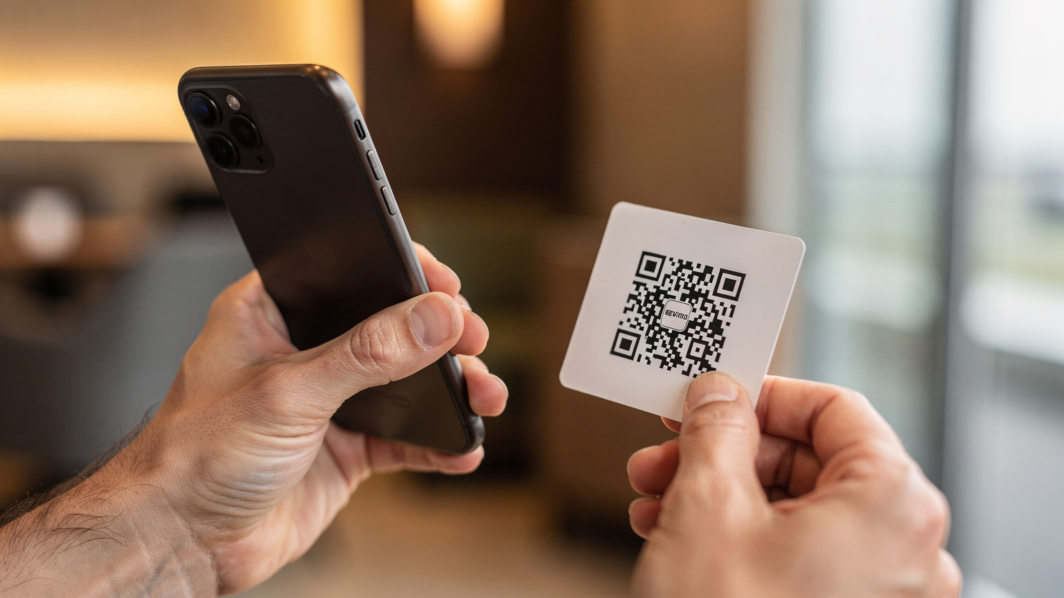 Zbliżenie na dłonie osoby trzymającej smartfon i skanującej kod QR w celu aktywacji eSIM, z niewyraźnym tłem hotelowego lobby lub lotniska.