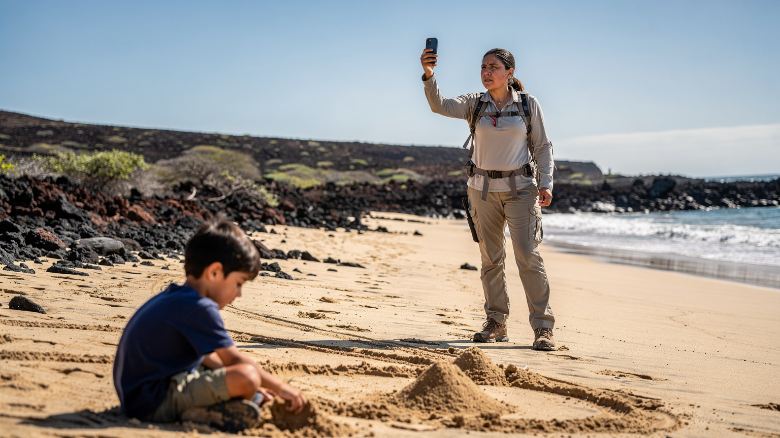 Uma mulher em Galápagos verificando a cobertura do celular enquanto seu filho brinca na areia, com uma paisagem vulcânica ao fundo.