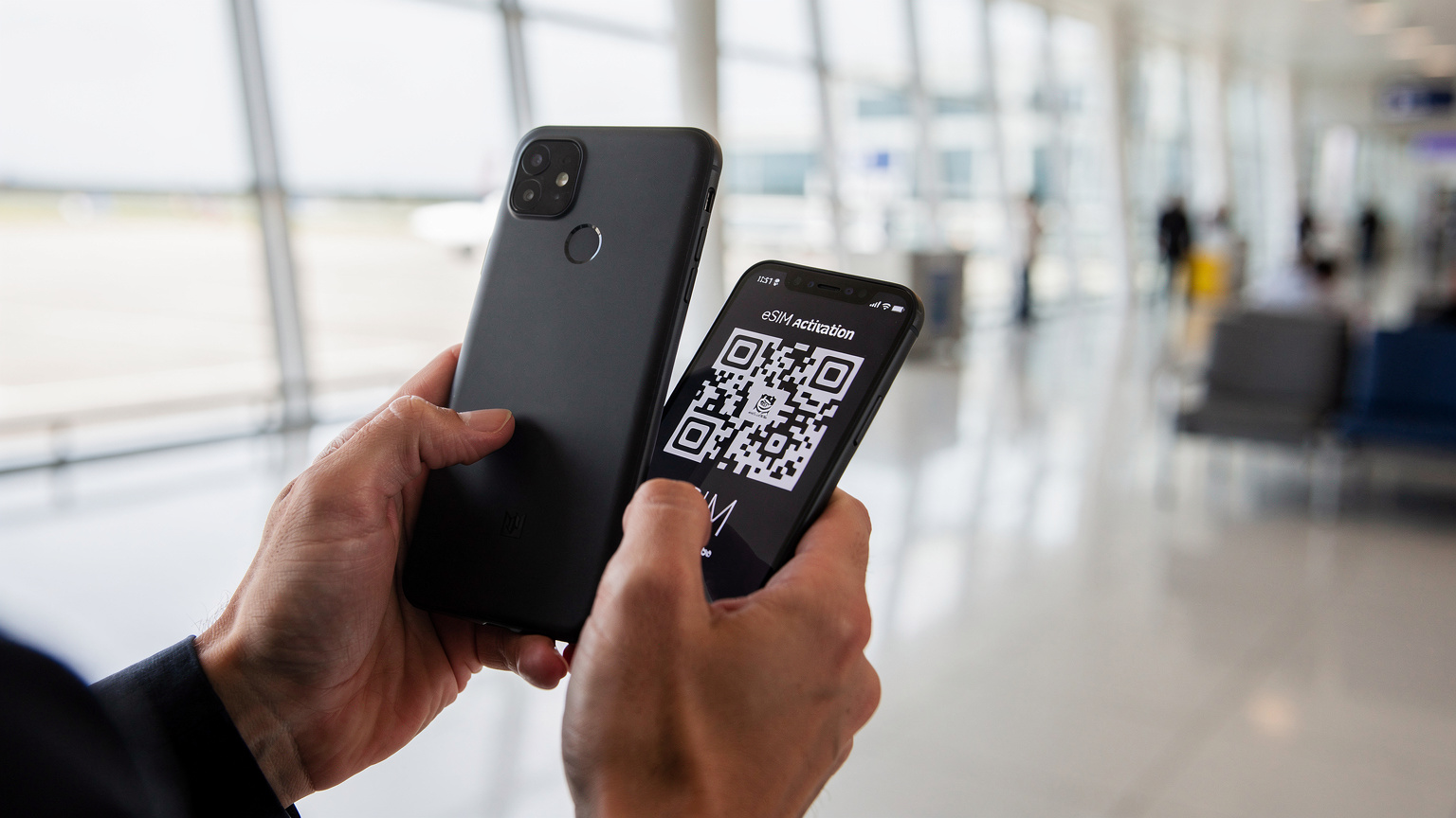 Tæt op af en persons hænder, der holder en smartphone op mod en anden smartphone, hvis skærm viser en QR-kode klar til scanning. Baggrunden er let sløret og viser en moderne lufthavnsterminal.