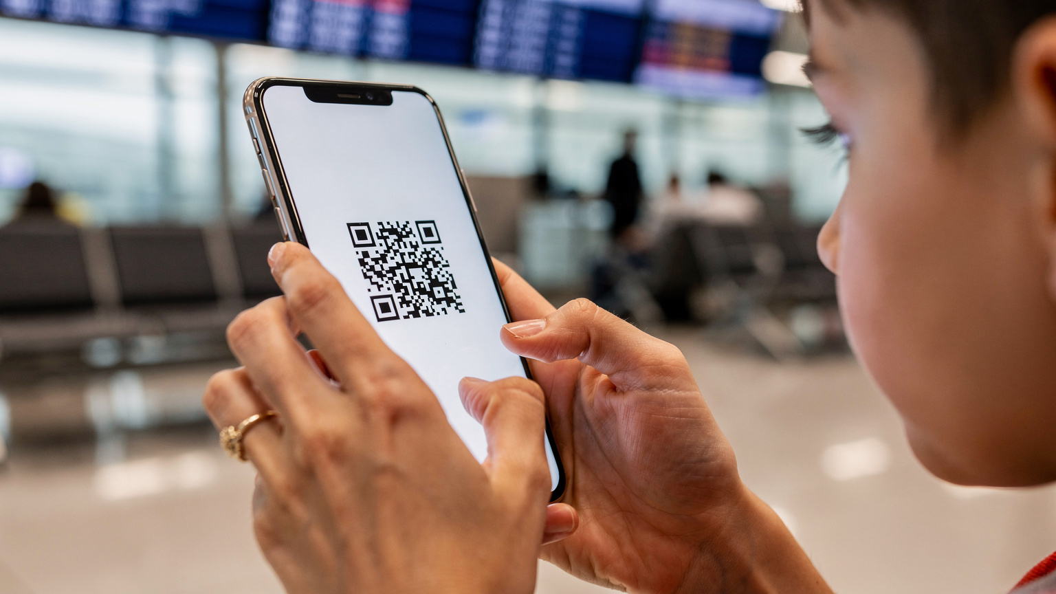 Zblízka záber na ženské ruky, ktoré držia smartfón a ukazujú dieťaťu na obrazovke proces aktivácie eSIM cez QR kód. V pozadí je rozmazané prostredie letiskovej haly s tabuľami odletov.