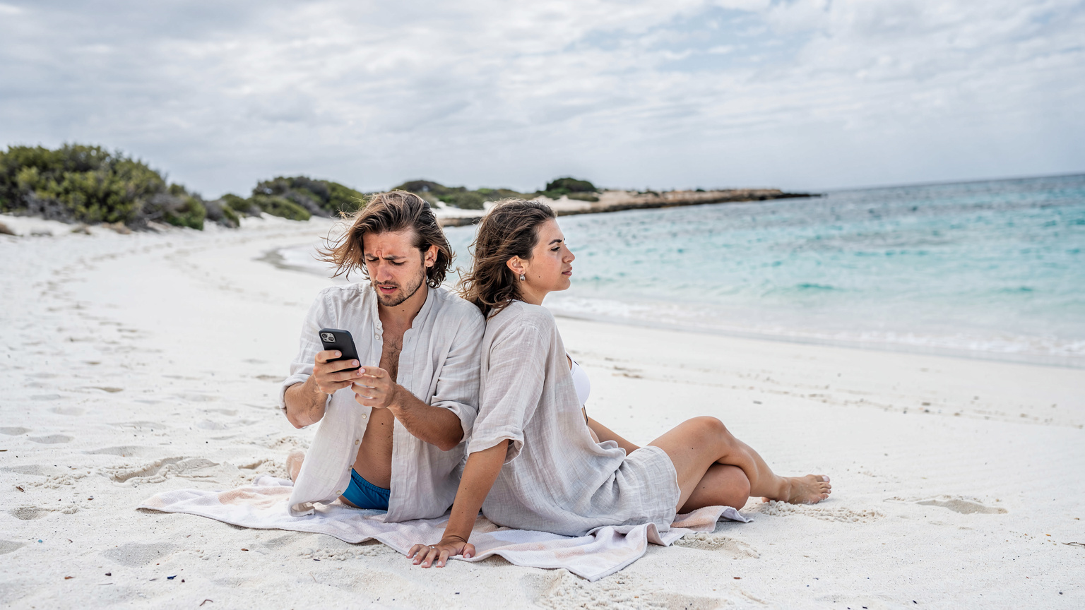 Una pareja joven comprueba la cobertura en su teléfono mientras se relajan en una hamaca en una tranquila playa de arena blanca en Formentera.