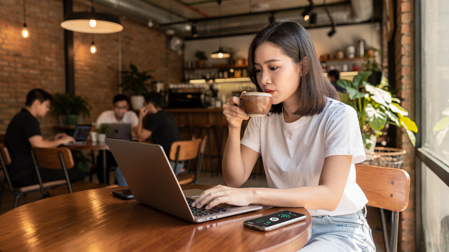 Một phụ nữ trẻ đang ngồi trong quán cà phê 'Work Saigon' ở Thành phố Hồ Chí Minh, vừa uống cà phê vừa làm việc trên máy tính xách tay, điện thoại thông minh đặt cạnh, hiển thị biểu tượng kết nối Wi-Fi và eSIM.