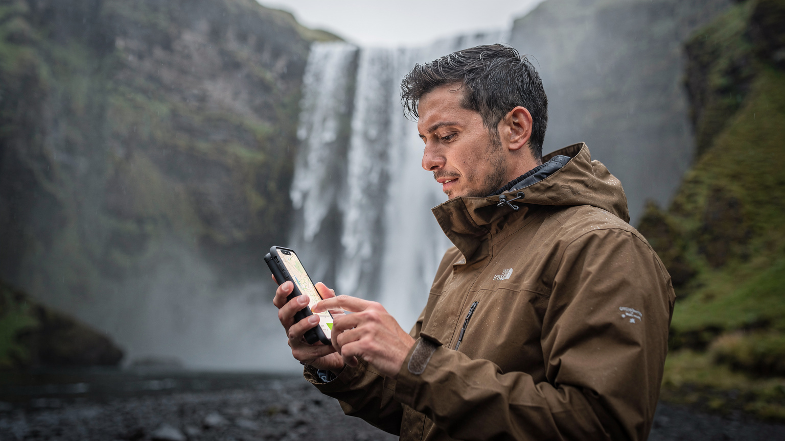 Cestovatel s telefonem v ruce kontroluje mapu u vodopádu Seljalandsfoss na Islandu, snaží se zachytit dokonalý okamžik.