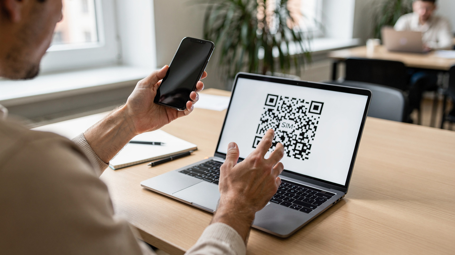 A close-up of a person's hands holding a smartphone, about to scan an eSIM QR code displayed on a laptop screen, with a blurry co-working space in the background.