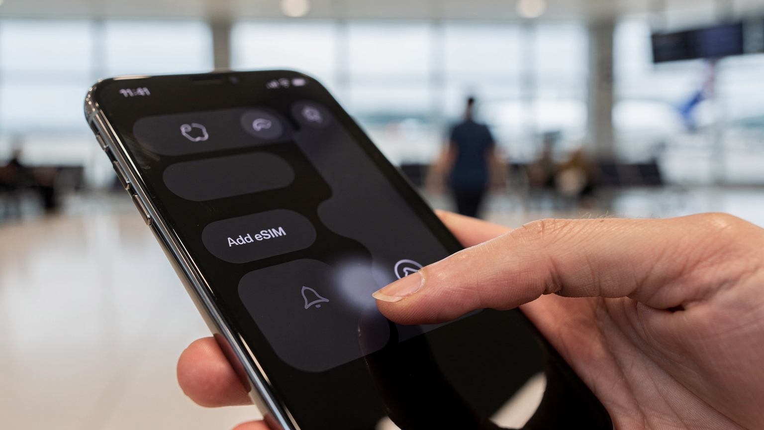 A close-up shot of an iPhone screen showing the 'Add eSIM' option in settings, with a finger about to tap it. The background is a gently blurred travel setting, perhaps an airport terminal.