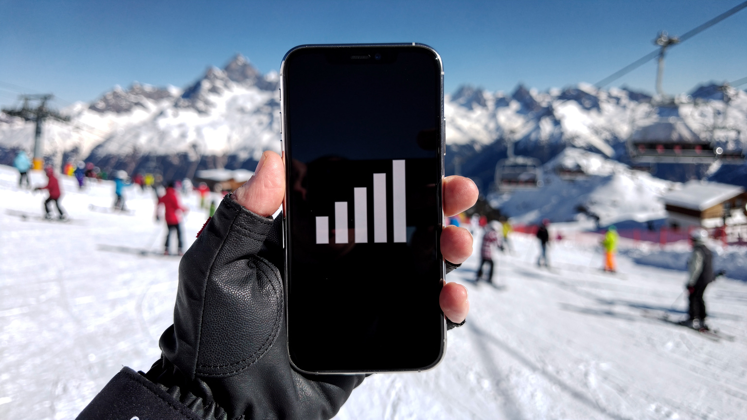Primer plano de una mano sosteniendo un teléfono con una señal de red fuerte, mientras de fondo se ve una pista de esquí en los Alpes suizos con esquiadores y montañas nevadas.