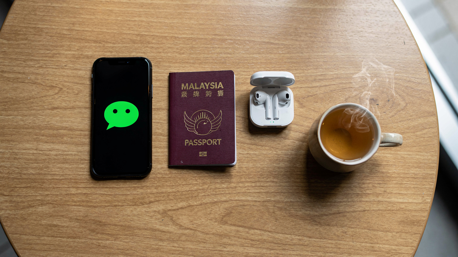 Sebuah flat-lay overhead sebuah meja kopi di kafe moden di Chengdu, memaparkan telefon pintar dengan aplikasi WeChat, pasport Malaysia, dan secawan teh jasmin.