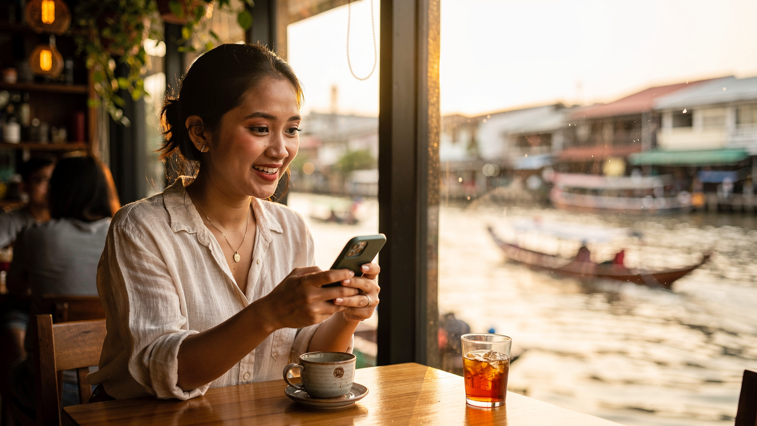 Seorang wanita tersenyum sambil memegang ponselnya di sebuah kafe tepi sungai yang ramai di tepi Sungai Chao Phraya, Bangkok, Thailand, dengan perahu-perahu tradisional melintas di latar belakang.