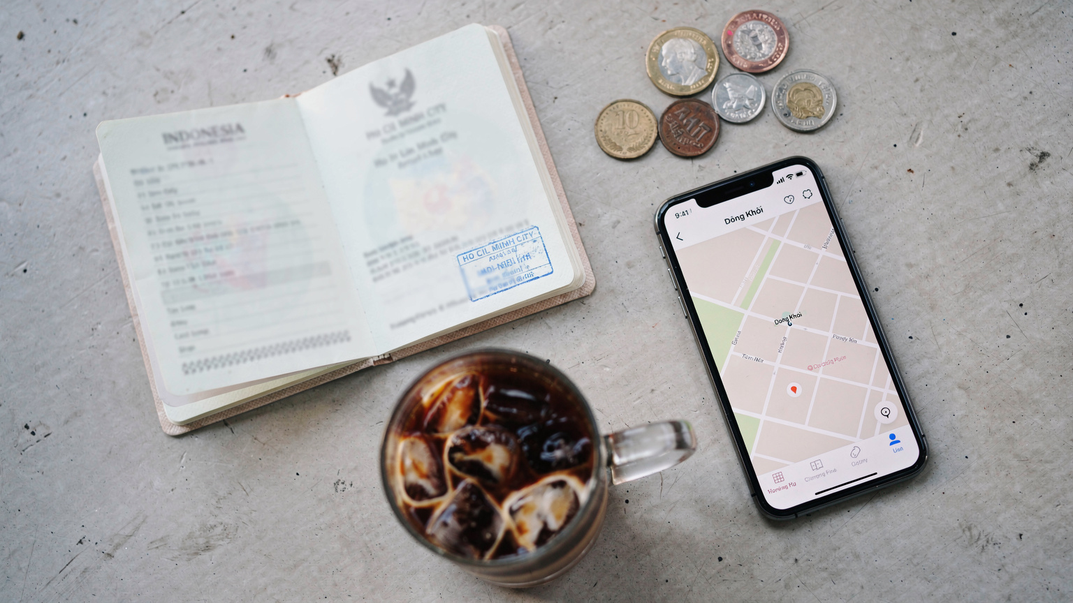 Tampilan overhead meja kafe dengan paspor Indonesia terbuka, ponsel cerdas yang menampilkan aplikasi peta, secangkir kopi, dan beberapa koin mata uang asing (contoh: Baht Thailand, Ringgit Malaysia) di sampingnya.