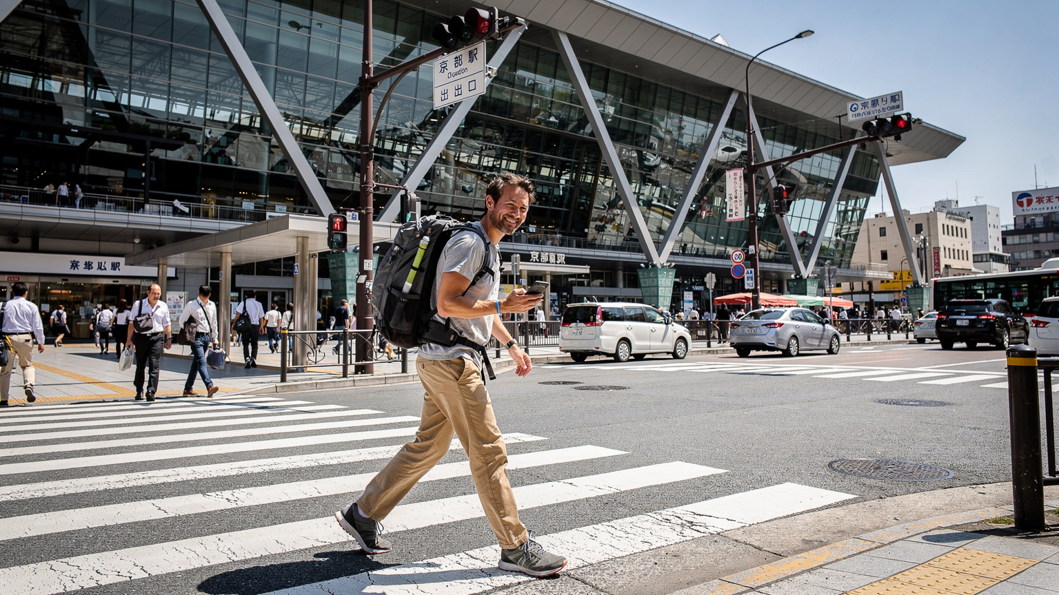 Un voyageur souriant, sac à dos et smartphone en main, traverse un passage piéton animé devant la gare de Kyoto, avec des panneaux de signalisation japonais en arrière-plan, symbolisant la navigation facile.